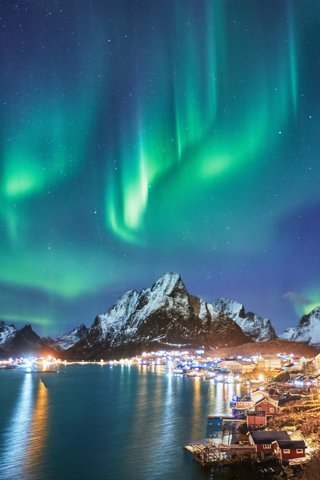 A view of a coastal town with a mountain backdrop, with the northern lights shiig green overhead