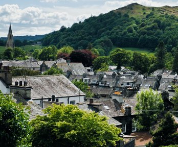 Rooftops of houses in a village with a church spire and hills with trees in the background. 