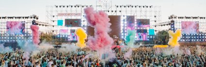 A view over a festival crowd with coloured smoke and bright TV screens
