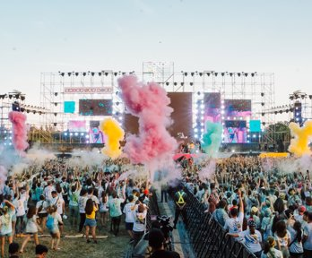 A view over a festival crowd with coloured smoke and bright TV screens