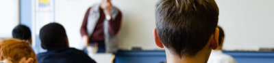 Several students faced towards a teacher at the front of a class room. The angle places one student in the foreground.