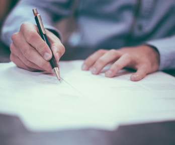 A person writes on a sheet of paper sticking out from a shallow stack of papers on top of a dark brown surface. The papers and background are blurred whist only the hands of the person can be seen