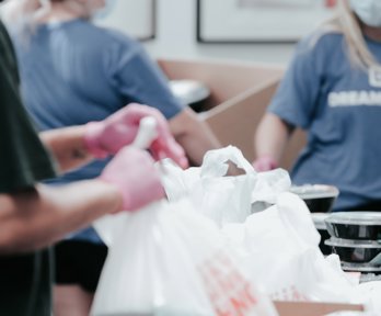 People in gloves handing out bags and pots of food. 