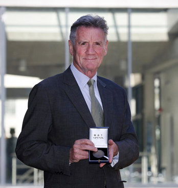 Michael Palin in a formal suit holding up a box with a medal inside.