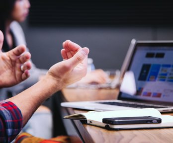 Person gesturing in a meeting with an open notebook, phone and laptop on a wooden table