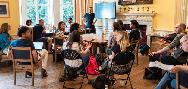 A presenter stands next to a screen with a presentation while listening attentively to an audience member talking.