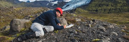 Researcher kneeling in a rocky landscape writing in a notebook. There is a glacier in the background.