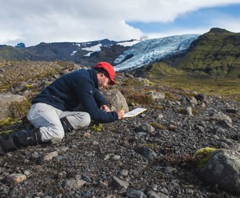 Researcher kneeling in a rocky landscape writing in a notebook. There is a glacier in the background.