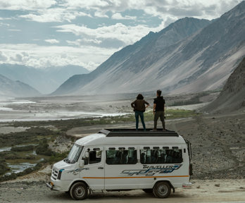 Two people are standing on top of a minibus, overlooking a dusty valley with a small river running along the bottom of it. There are mountains to the right hand side and the sky is cloudy.