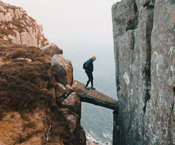 A person is standing on a precariously placed rock that is wedged between two cliff sides. They are high up and there is a long drop beneath the wedged rock onto a rocky beach.