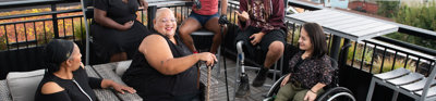 Six people sit down in chairs on a porch laughing. One person sits in a wheel chair, another holds a cain and another has a prosthetic leg.