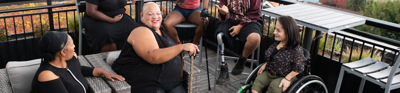 Six people sit down in chairs on a porch laughing. One person sits in a wheel chair, another holds a cain and another has a prosthetic leg.