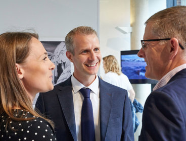 Three people in professional attire smiling and talking while standing in a bright exhibition space.