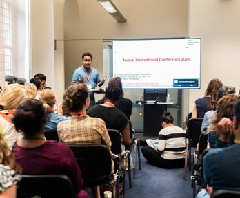 A presenter speaks to a room of people. A large screen show a slide of a PowerPoint presentation.