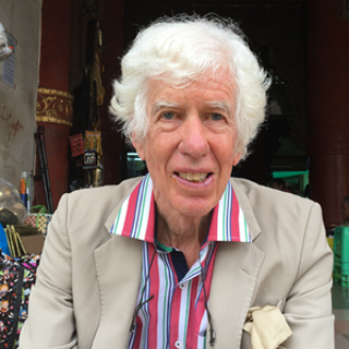 A man with white hair and wearing a colourful stripped shirt with a formal, beige jacket sitting outside a pagoda.