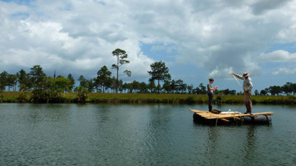 Two people stand on a wooden raft  in the middle of a lake. The lake is surrounded by green grass land, sparsely dotted with trees.