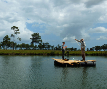 Two people stand on a wooden raft  in the middle of a lake. The lake is surrounded by green grass land, sparsely dotted with trees.